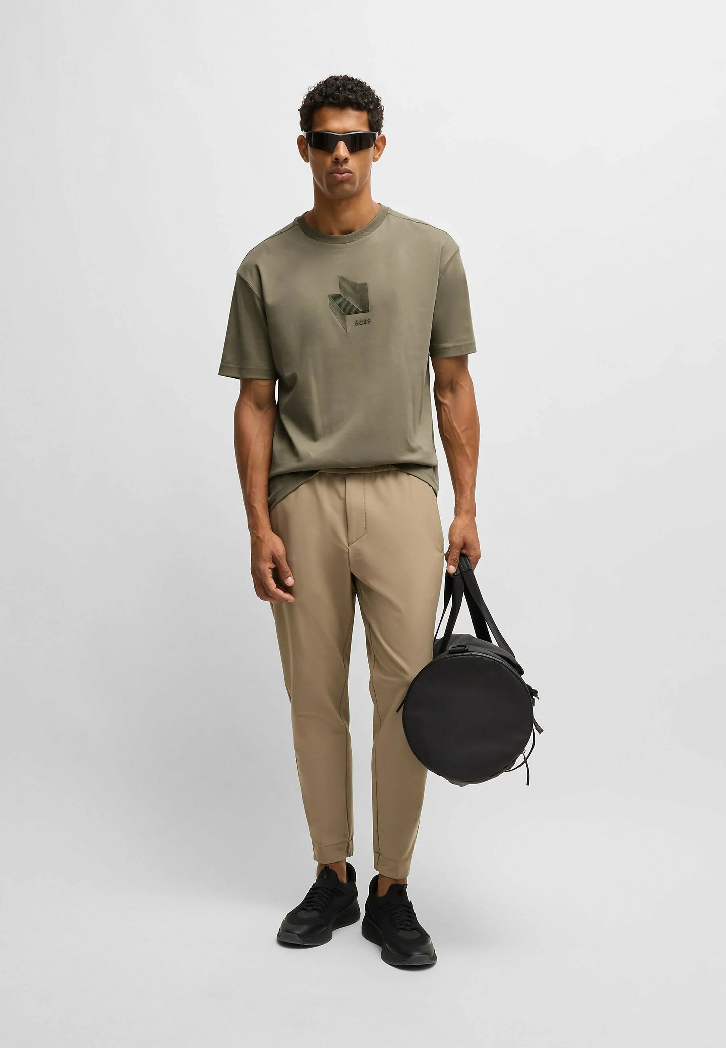 Man holding a black bag wearing a green t-shirt and beige pants on a white background